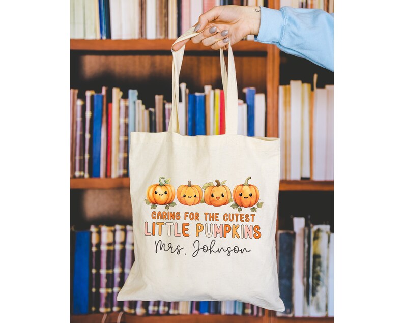 K&ouml;nnte beinhalten: Eine wei&szlig;e Stofftasche mit einem Design aus f&uuml;nf orangefarbenen K&uuml;rbissen mit l&auml;chelnden Gesichtern. Der Text auf der Tasche lautet "Caring for the cutest little pumpkins Mrs. Johnson".