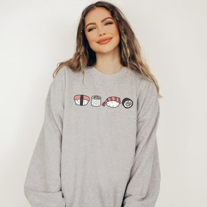 May include: A gray sweatshirt with a graphic of five different sushi rolls. The rolls are drawn in a cartoon style and include salmon, shrimp, and avocado.