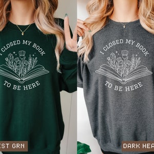 K&ouml;nnte beinhalten: Ein gr&uuml;nes und ein dunkel-heather-graues Sweatshirt mit einem wei&szlig;en Grafikdesign eines offenen Buches, aus dem Blumen wachsen. Der Text auf dem Sweatshirt lautet "Ich habe mein Buch geschlossen, um hier zu sein".