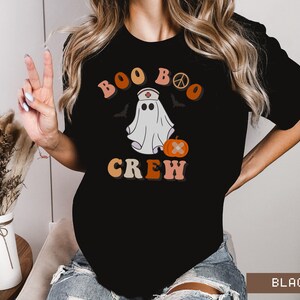 May include: Black t-shirt with a retro-style graphic of a ghost wearing a nurse's hat and holding a pumpkin. The text "Boo Boo Crew" is written in a colorful, retro font.