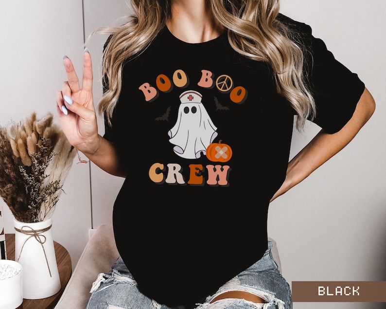 May include: Black t-shirt with a retro-style graphic of a ghost wearing a nurse's hat and holding a pumpkin. The text "Boo Boo Crew" is written in a colorful, retro font.
