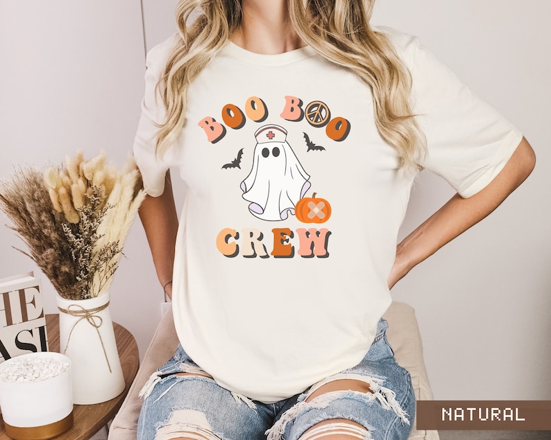 May include: White t-shirt with a graphic of a ghost wearing a nurse's hat and holding a pumpkin. The text "Boo Boo Crew" is written in a retro font with a peace sign.