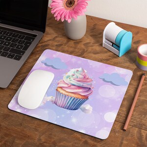 May include: A purple and blue mouse pad with a white computer mouse on top. The mouse pad features a whimsical illustration of a pink and blue cupcake with sprinkles and a swirl of frosting. The cupcake is surrounded by white clouds and a light purple background with white dots.
