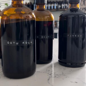 May include: Three amber glass bottles with black lids, each containing a dark liquid. The bottles are labeled "GOTU KOLA", "N GRAPE", and "YARROW". The bottles are on a white countertop.