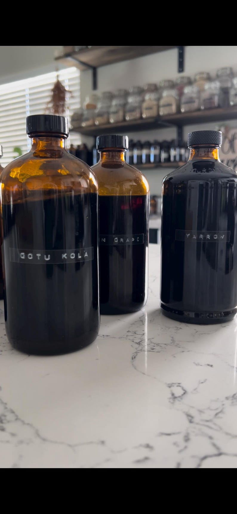 May include: Three amber glass bottles with black lids, each containing a dark liquid. The bottles are labeled "GOTU KOLA", "N GRAPE", and "YARROW". The bottles are on a white countertop.