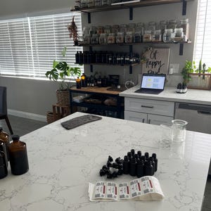 May include: A kitchen scene with a white countertop and various apothecary supplies. There are rows of small black bottles, labels, and glass beakers. Shelves hold numerous glass jars filled with herbs and spices. A laptop displays a presentation, and a sign reads "Off Happy Day."