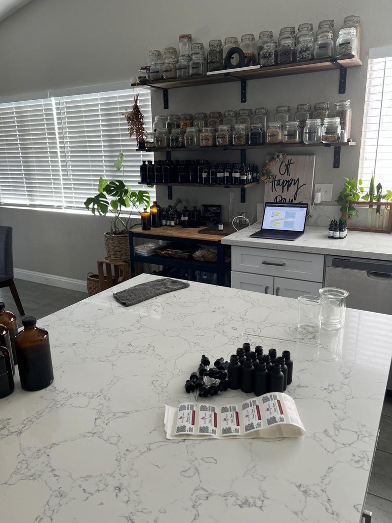 May include: A kitchen scene with a white countertop and various apothecary supplies. There are rows of small black bottles, labels, and glass beakers. Shelves hold numerous glass jars filled with herbs and spices. A laptop displays a presentation, and a sign reads "Off Happy Day."