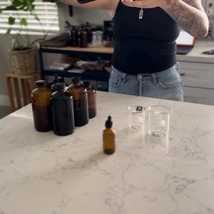 May include: A person pouring liquid from a brown glass bottle into a small glass beaker. Several brown glass bottles and a dropper bottle are on the counter. The scene suggests a laboratory or apothecary setting.