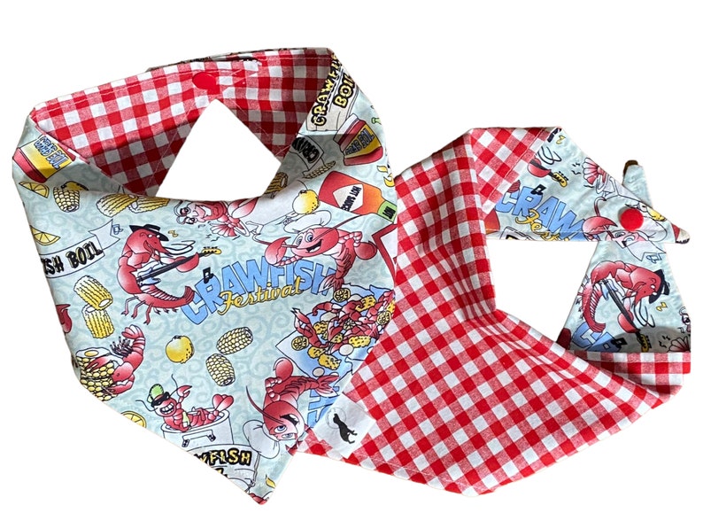 May include: A red and white checkered bandana with a blue and white print featuring crawfish, corn, and lemons. The print includes the text "Crawfish Festival".