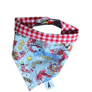 May include: A blue and white bandana with a red and white checkered border. The bandana features a cartoon illustration of crawfish, corn on the cob, and the text "Crawfish Festival".