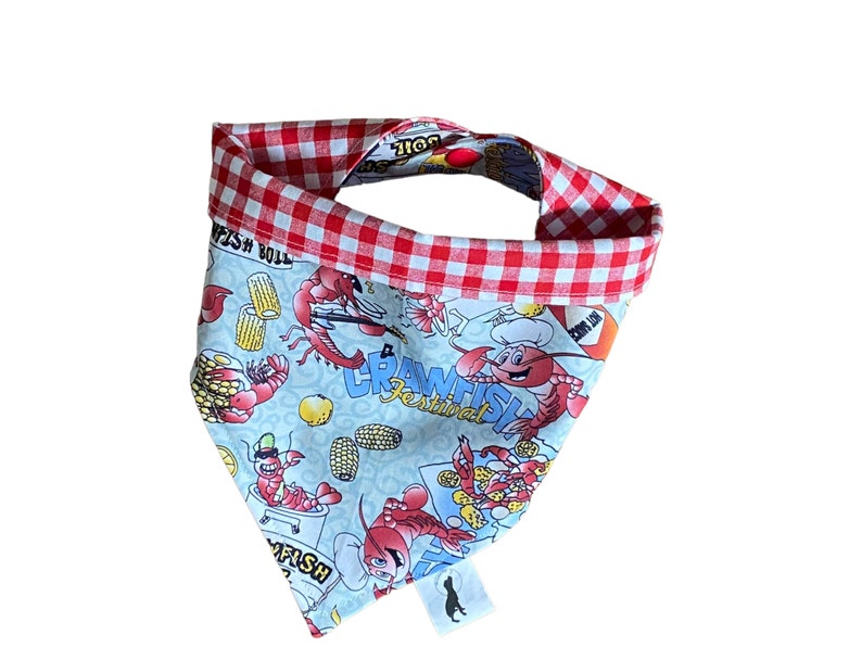 May include: A blue and white bandana with a red and white checkered border. The bandana features a cartoon illustration of crawfish, corn on the cob, and the text "Crawfish Festival".