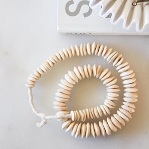 May include: A coiled strand of light-colored wooden beads with a natural finish. The beads are strung on a cream-colored cord. A white book with the word "STUDIO" is visible, along with white paper decorations and bronze bell-shaped ornaments.