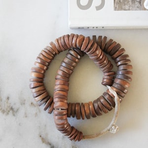 May include: A necklace made of stacked, round, wooden beads in shades of brown and tan. The beads are strung together with a light-colored cord. A white book with the word "STILL" is in the background.