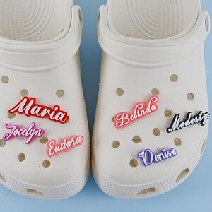 May include: Two white clogs with various coloured personalised name charms. The charms spell out the names Maria, Jaclyn, Eudora, Belinda, Denise, and Modesty.
