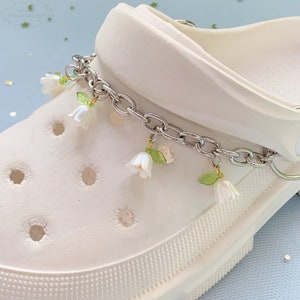 May include: White clog shoe with a silver chain and white flower charms. The chain has small green leaves attached to the charms.