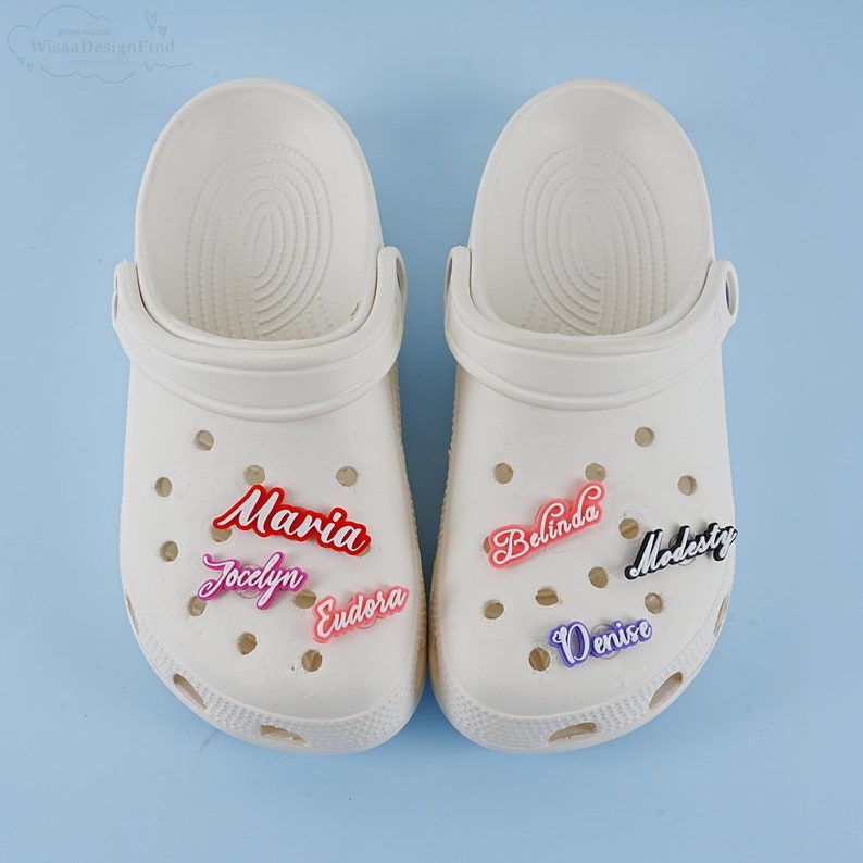 May include: Two white clogs with various coloured personalised name charms. The charms spell out the names Maria, Jaclyn, Eudora, Belinda, Denise, and Modesty.