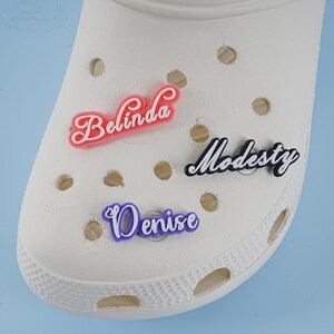 May include: Three personalised shoe charms with the names Belinda, Modesty, and Denise. The charms are made of a soft, flexible material and are in pink, black, and purple.