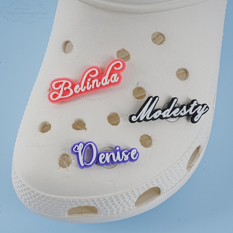 May include: Three personalised shoe charms with the names Belinda, Modesty, and Denise. The charms are made of a soft, flexible material and are in pink, black, and purple.