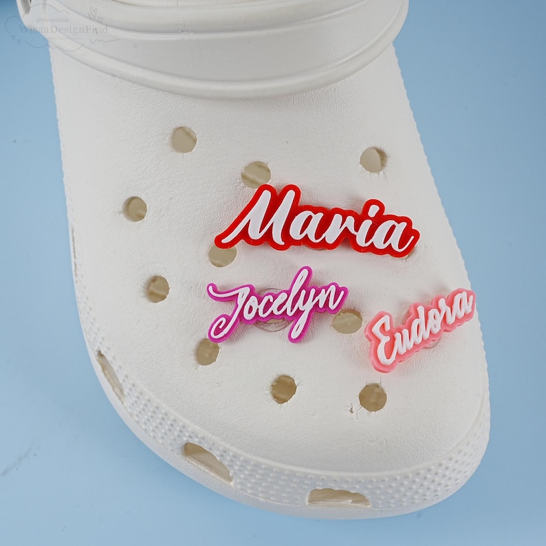 May include: Three personalised name charms in red, pink and purple. The charms are attached to a white clog shoe. The names are Maria, Jocelyn and Eudora.