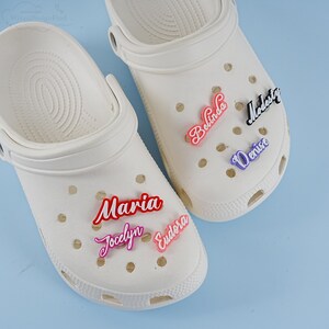 May include: Two white clogs with colourful personalised name charms. The charms spell out "Maria", "Jocelyn", "Eudora", "Belinda", "Madeste", and "Denise".