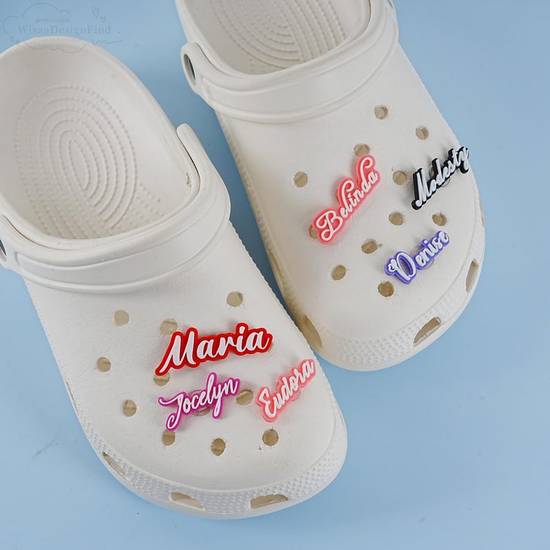 May include: Two white clogs with colourful personalised name charms. The charms spell out "Maria", "Jocelyn", "Eudora", "Belinda", "Madeste", and "Denise".