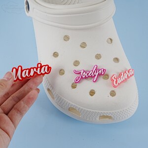 May include: White clogs with three personalised name tags. The name tags are red, pink and light pink. The names on the tags are Maria, Jocelyn and Eudora.