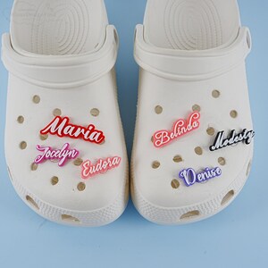 May include: Two white clogs with colourful personalised name charms. The charms spell out the names Maria, Jocelyn, Eudora, Belinda, Denise, and Modesty.