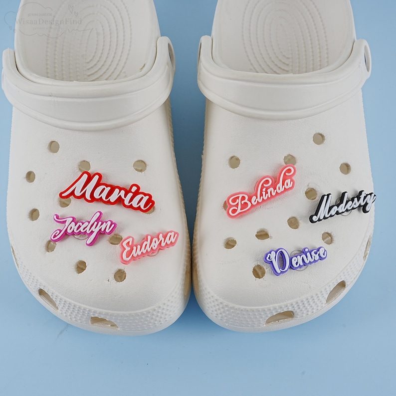 May include: Two white clogs with colourful personalised name charms. The charms spell out the names Maria, Jocelyn, Eudora, Belinda, Denise, and Modesty.