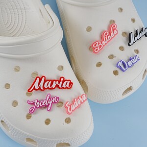 May include: Two white clogs with colourful personalised name charms. The charms spell out the names Maria, Jocelyn, Eudora, Belinda, Denise, and Modesty.