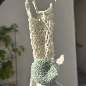 Könnte beinhalten: Handgefertigte Häkeltasche in Form einer Schildkröte. Die Tasche hat einen cremefarbenen, offen gewebten Körper und einen grünen Schildkrötenpanzer. Die Schildkröte hat weiße Flossen und einen Kopf.