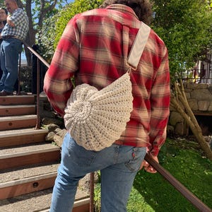 Op de afbeelding: Een beige gehaakte schoudertas met een cirkelvormig ontwerp aan de voorkant. De tas heeft een crèmekleurige band met een zigzagpatroon. De persoon draagt een rood en tan geruit overhemd en een blauwe jeans.