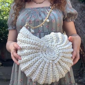 Peut inclure: Un sac &agrave; main en crochet de couleur cr&egrave;me avec un motif en spirale. Le sac est dot&eacute; d'une bandouli&egrave;re en cha&icirc;ne dor&eacute;e orn&eacute;e de fausses perles. Le sac est tenu devant une personne portant une robe &agrave; fleurs.