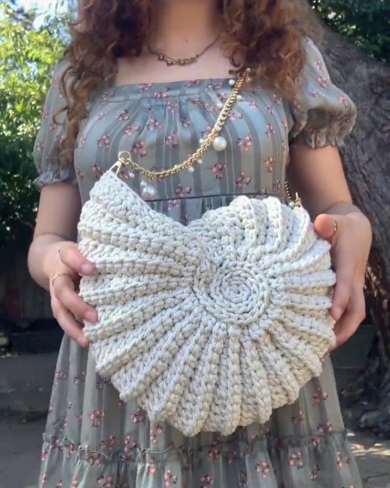 Peut inclure: Un sac &agrave; main en crochet de couleur cr&egrave;me avec un motif en spirale. Le sac est dot&eacute; d'une bandouli&egrave;re en cha&icirc;ne dor&eacute;e orn&eacute;e de fausses perles. Le sac est tenu devant une personne portant une robe &agrave; fleurs.