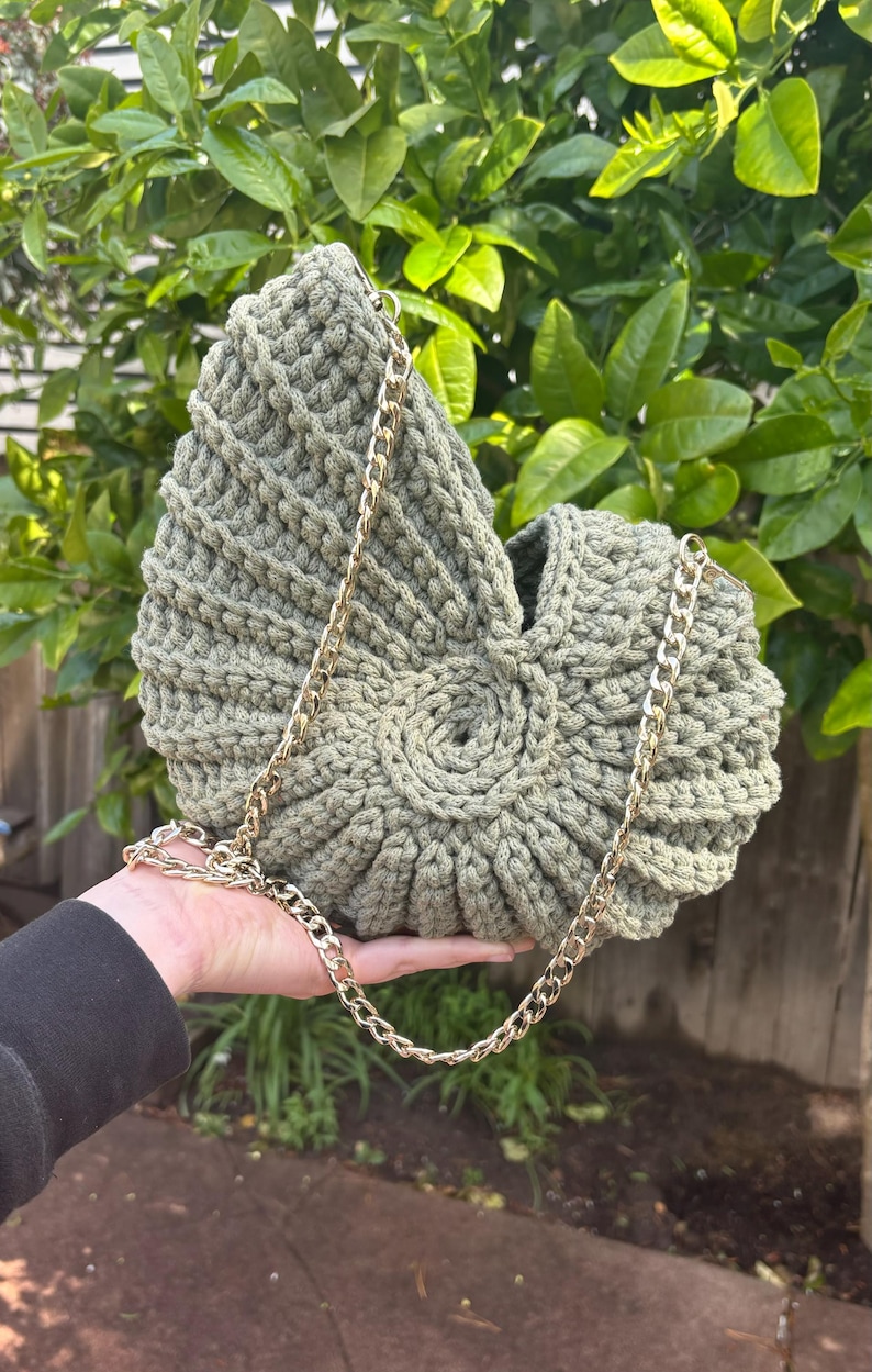 Peut inclure: Sac &agrave; main en crochet vert olive avec une cha&icirc;ne dor&eacute;e. Le sac pr&eacute;sente un motif en spirale sur le devant et un motif tiss&eacute; textur&eacute;. La cha&icirc;ne est dor&eacute;e et le sac est tenu dans une main.