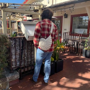 Peut inclure: Une personne portant une chemise &agrave; carreaux rouge et noir et un jean bleu, portant un sac en crochet blanc avec une sangle cr&egrave;me. Le sac a un design en forme d'&eacute;ventail. La personne est &agrave; l'ext&eacute;rieur d'un b&acirc;timent avec une enseigne.