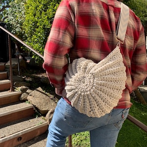 Peut inclure: Un sac &agrave; bandouli&egrave;re beige en crochet avec un motif en spirale. Le sac a une sangle &agrave; motifs et est port&eacute; sur une chemise &agrave; carreaux rouge et beige et un jean bleu. L'arri&egrave;re-plan comprend un feuillage vert et un escalier.
