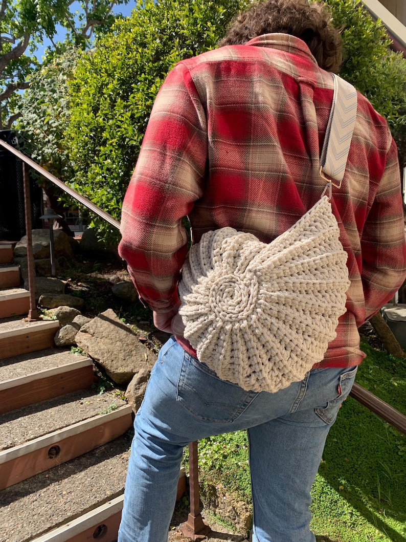 Peut inclure: Un sac &agrave; bandouli&egrave;re beige en crochet avec un motif en spirale. Le sac a une sangle &agrave; motifs et est port&eacute; sur une chemise &agrave; carreaux rouge et beige et un jean bleu. L'arri&egrave;re-plan comprend un feuillage vert et un escalier.