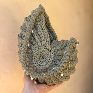 Peut inclure: Un panier crochet&eacute; vert sauge avec un motif en spirale sur le devant. Le panier est orn&eacute; de breloques dor&eacute;es et de fausses perles pendantes. Le panier fait main est tenu dans la main d'une personne.
