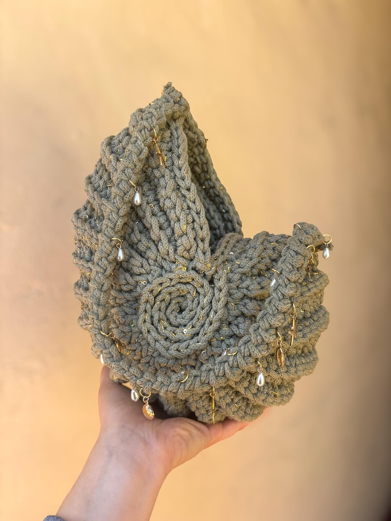 Peut inclure: Un panier crochet&eacute; vert sauge avec un motif en spirale sur le devant. Le panier est orn&eacute; de breloques dor&eacute;es et de fausses perles pendantes. Le panier fait main est tenu dans la main d'une personne.