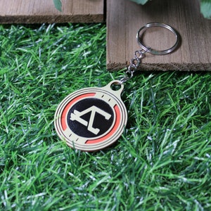 May include: A round keychain with a silver chain and ring. The keychain has a cream-colored border, a red inner ring, and a black center with a white geometric design. The keychain is on a bed of green grass.