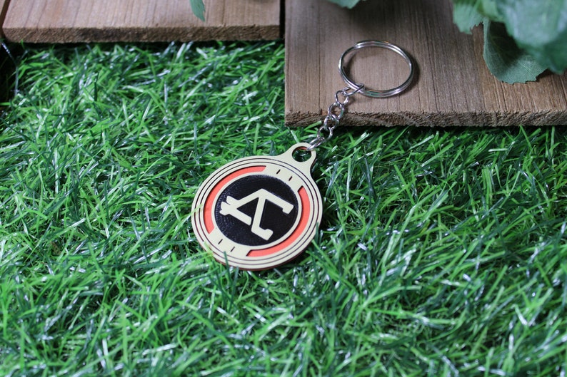 May include: A round keychain with a silver chain and ring. The keychain has a cream-colored border, a red inner ring, and a black center with a white geometric design. The keychain is on a bed of green grass.