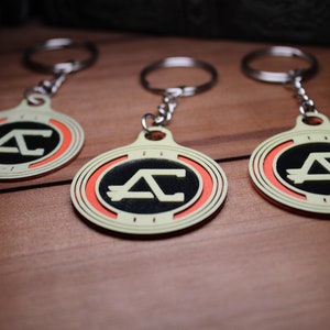 May include: Three keychains with a circular design featuring a black center with a white "AC" logo, surrounded by an orange ring and a cream-colored border. Each keychain has a silver keyring attached.