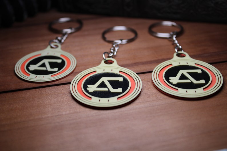 May include: Three keychains with a circular design featuring a black center with a white "AC" logo, surrounded by an orange ring and a cream-colored border. Each keychain has a silver keyring attached.