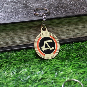 May include: A round keychain with a gold-colored border and a black center featuring a stylized letter "A" in white. The keychain has a red ring around the black center. The background includes a dark book and green grass.