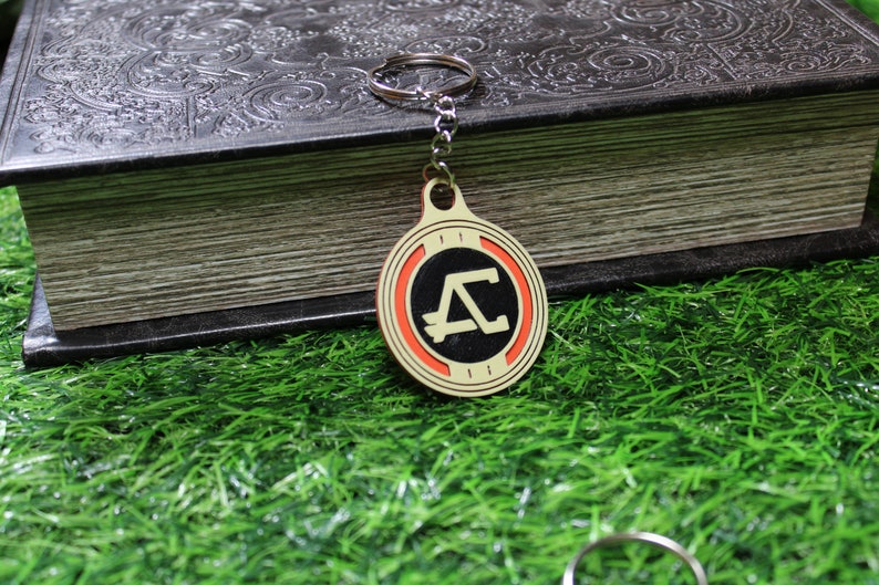 May include: A round keychain with a gold-colored border and a black center featuring a stylized letter "A" in white. The keychain has a red ring around the black center. The background includes a dark book and green grass.