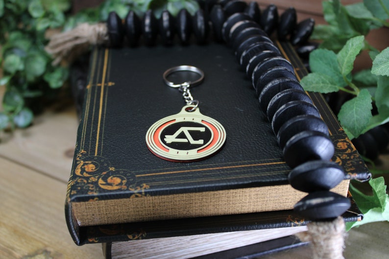 May include: A gold-colored keychain with a red and cream-colored circular design featuring the letters "AC". The keychain hangs from a silver ring. The keychain rests on a vintage book with gold accents, next to a strand of black beads.