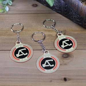May include: Three keychains with a circular design featuring a black letter "A" and a "C" inside a black circle, bordered by orange and cream. Each keychain has a silver ring and chain. The keychains are on a wooden surface.