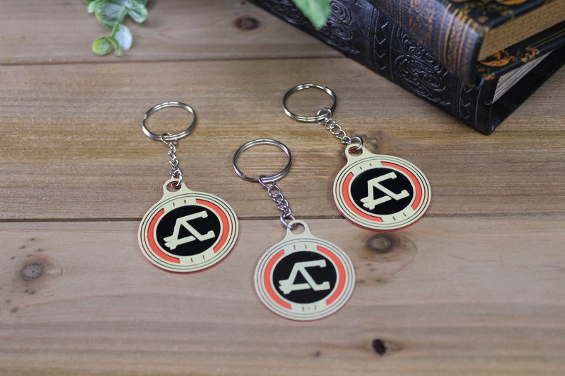 May include: Three keychains with a circular design featuring a black letter "A" and a "C" inside a black circle, bordered by orange and cream. Each keychain has a silver ring and chain. The keychains are on a wooden surface.