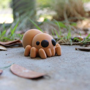 Puede incluir: Una pequeña figura de araña de color marrón impresa en 3D con ojos negros. La araña está sentada sobre una superficie de hormigón gris.