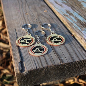 May include: Three keychains with a gold-colored circular pendant featuring a black letter "A" inside a red and gold border. The keychains are on a wooden surface.